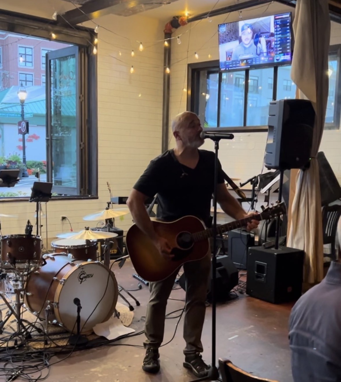 Johnny Torres singing into a microphone while playing an acoustic guitar in a small venue with string lights and a drum kit behind him.