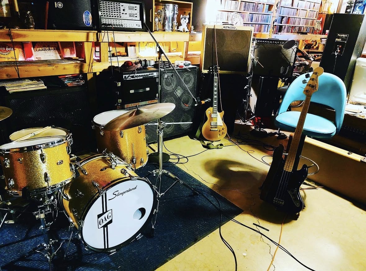 Drum kit set up in a rehearsal room with amps, guitars, and a bass leaning nearby.