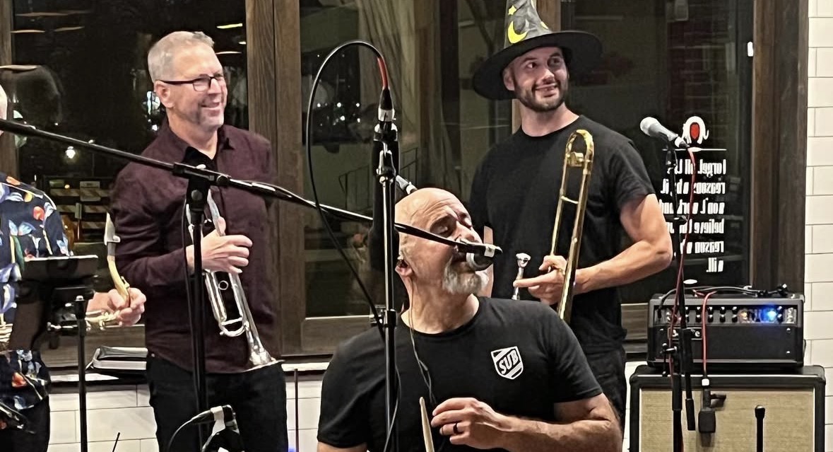 Slackers Unite Band performing indoors with Johnny Torres at the microphone while bandmates hold a trumpet and trombone.
