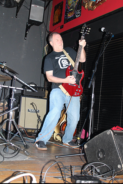 Dave Lysein playing a red electric guitar on a small indoor stage, wearing a black T-shirt and jeans, with amplifiers, drum kit, and microphone stand visible around him.