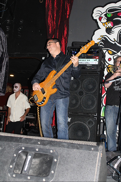 Mike Lahart playing a natural wood electric bass guitar on a small indoor stage, standing in front of stacked speakers and audio equipment, with other band members partially visible.