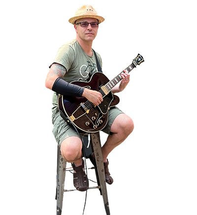Vito Masotti,  wearing a straw hat and glasses seated on a metal stool, holding a dark hollow-body electric guitar against a plain light background.