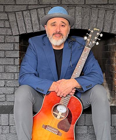 Man wearing a blue blazer and light blue hat sitting in front of a brick fireplace holding a sunburst Gibson acoustic guitar.
