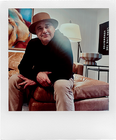Man wearing a brown hat and black sweater sitting on a leather couch in a living room, presented in a Polaroid-style photo frame.